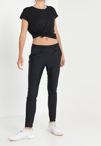 Czarna koszulka z węzełkiem i dopasowane czarne spodnie. Spodnie mają boczne kieszenie, gładką teksturę i zwężający się kształt, w połączeniu z białymi trampkami.