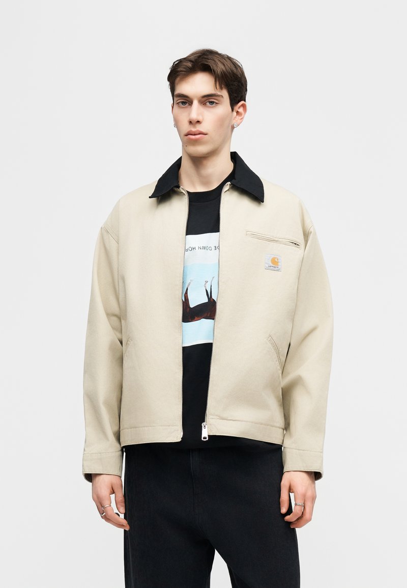 Joven con chaqueta beige con cierre y cuello negro, camiseta negra con gráfico y pantalones negros, de pie frente a un fondo liso.