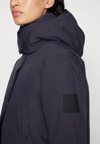 Giacca outdoor blu navy con una superficie texturizzata, colletto alto, cappuccio staccabile e un'etichetta quadrata nera del marchio sulla manica.