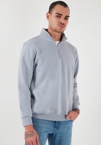 Sudadera de cremallera gris claro en tejido suave, con cuello alto y mangas largas, puños y dobladillo acanalados, combinada con jeans azules.