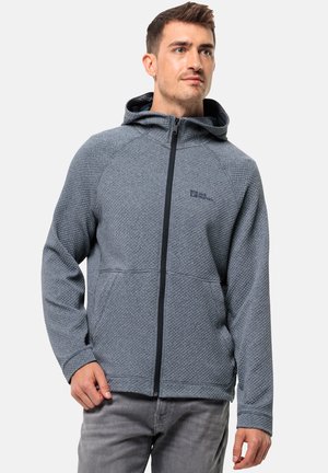 Hombre que lleva una chaqueta con capucha de textura gris y cremallera, con bolsillos, de pie contra un fondo claro y liso.