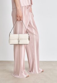 Helle rosa plissierte Hosen mit einem passenden Oberteil, kombiniert mit einer cremefarbenen Handtasche mit strukturiertem Design und metallischen Akzenten.