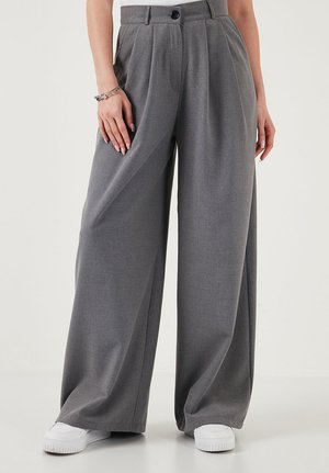 Hallid laiade säärtega püksid, voldid, nööbitav vöökoht, kantud valgete tenniste ja vasakul randmel olevale käevõruga.