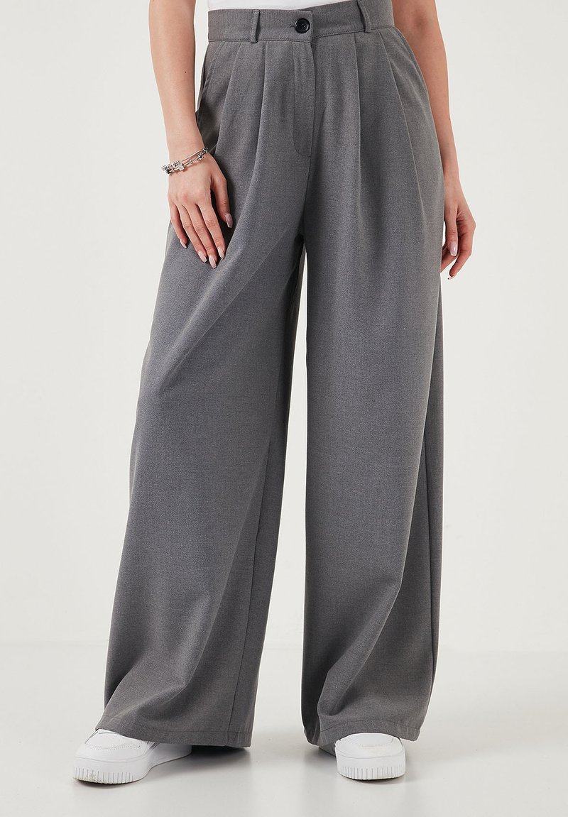 Wide-leg gray trousers with pleats, buttoned waist, worn with white sneakers and a bracelet on the left wrist.