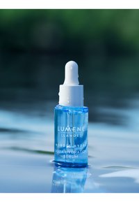 Klar glasdråberflaske med en hvid låg, der indeholder blåt serum. Etiketten har hvid tekst. Baggrunden er en blødt sløret vandoverflade.
