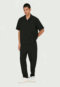 Camisa preta de mangas curtas com bolso, combinada com calças a condizer e ténis brancos. Textura suave, ajuste descontraído, design minimalista.