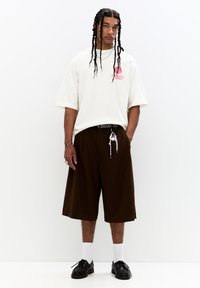 Oversized cream t-shirt with red text, paired with brown knee-length shorts. Black shoes, white socks, and accessories include a chain and bracelet.