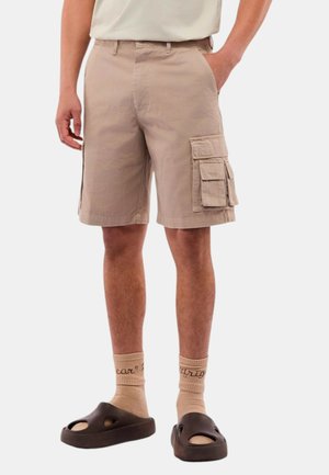 Pantaloni cargo beige con tasche laterali, zip e vestibilità regolare. Indossati con sandali scivolati marroni e calze beige.
