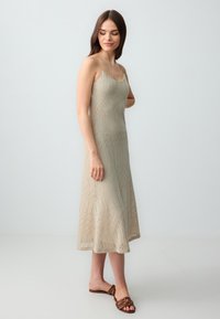 Beige, gebreide jurk tot op de knie met spaghettibandjes, voorzien van een zigzagpatroon en een licht uitlopende zoom. Gepaarde met bruine sandalen.