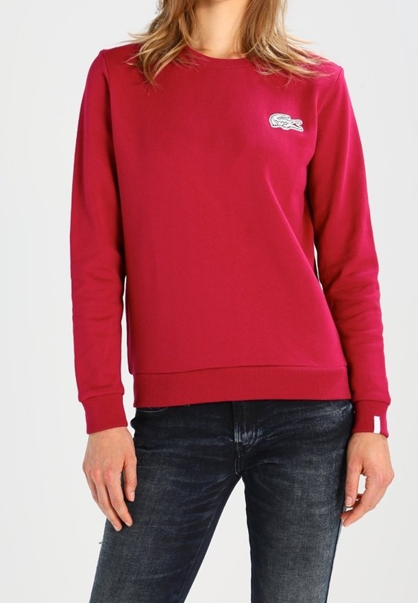 Personne portant un sweat-shirt rouge à manches longues avec un petit logo blanc sur la poitrine et un jean bleu foncé, debout devant un fond uni.