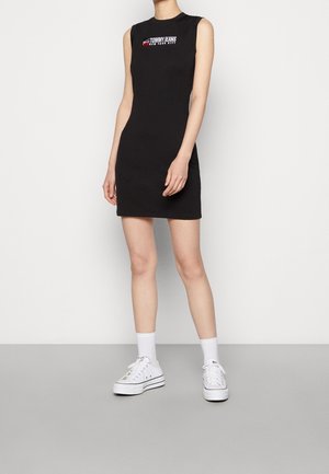 Mujer con vestido sin mangas negro de Tommy Jeans con logo, calcetines blancos y zapatillas blancas de plataforma, posando frente a un fondo liso.