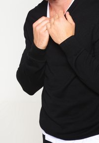 Zwarte v-hals sweater met lange mouwen, een gladde textuur, een aansluitend ontwerp en een wit onderhemd zichtbaar bij de halslijn.