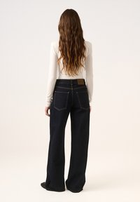 Jeans w szerokich nogawkach z ciemnego denimu z wysokim stanem, kontrastowym żółtym przeszyciem i klasycznymi tylnymi kieszeniami. W połączeniu z dopasowanym białym topem z długim rękawem.