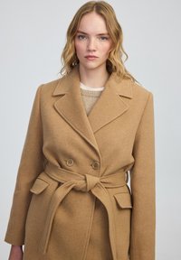 Manteau en laine camel avec un design croisé, des revers crantés, des poches latérales et une ceinture à nouer à la taille. Texture lisse, couleur chaude.