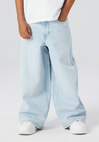 Gaiši zilas, platas džinsa bikses ar klasisku piecu kabatu dizainu un nodilušu apdari, kopā ar baltiem sporta apaviem.