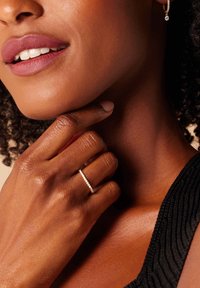 Zilveren ring met kleine heldere stenen, gedragen aan een hand met glanzende huid. Textuur van zwarte kleding en natuurlijke krullen zijn zichtbaar op de achtergrond.