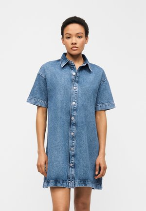 Frau in einem kurzärmeligen Jeanskleid mit Knopfleiste und Kragen, die vor einem schlichten hellen Hintergrund steht.