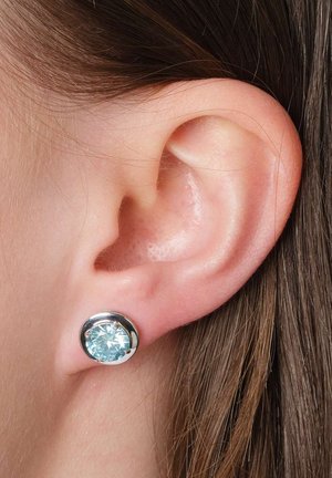 Nahaufnahme eines Ohrs einer Person mit braunem Haar, die einen silbernen Steckohrring mit einem hellblauen Edelstein trägt.