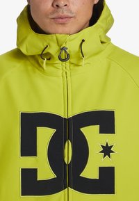 Ljust limegrön vattentät jacka med en svart DC-logotyp på framsidan. Har en huva med snoddar och en slät, matt yta.