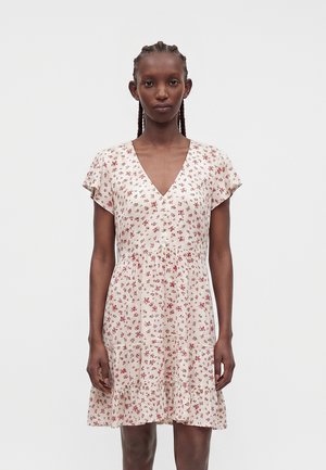 Žena s vrkočmi, ktorá nosí krémové šaty s krátkym rukávom a výstrihom do V s červeným kvetinovým vzorom, stojaca na jednoduchom bielom pozadí.