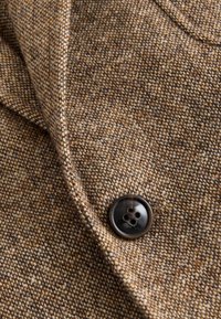 Tissu en tweed brun avec un motif texturé, arborant un bouton sombre à quatre trous. Le tissu met en valeur différentes nuances de brun.