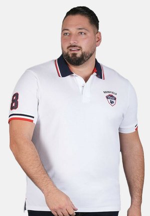 Homme portant un polo blanc avec un col et des poignets de manches rayés bleu marine et rouge, debout avec une expression neutre, regardant vers la droite.