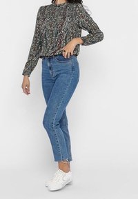 Bluzka z długim rękawem w wielokolorowym materiale z teksturowanym wzorem, w komplecie z niebieskimi dżinsami o wysokim stanie i prostymi nogawkami oraz białymi trampkami.