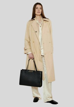 Beige trenchcoat med dobbeltknapdesign, guldknapper, parret med en cremefarvet satinbluse og mønstrede bukser. Sort håndtaske med kædestropper.