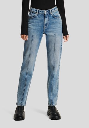 Femme portant un jean taille haute bleu clair avec des coutures verticales à l'avant, un haut noir à manches longues et des bottes épaisses noires, debout sur un fond gris.