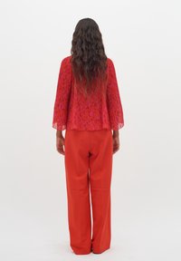 Blouse transparente à motifs rouges avec manches évasées, associée à un pantalon rouge à jambes larges. Tissu lisse avec un design fluide, détail visible dans le dos.