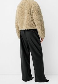 Beige faux fur-jakke med en tekstureret finish, parret med sorte faux læder brede bukser med elastisk talje.