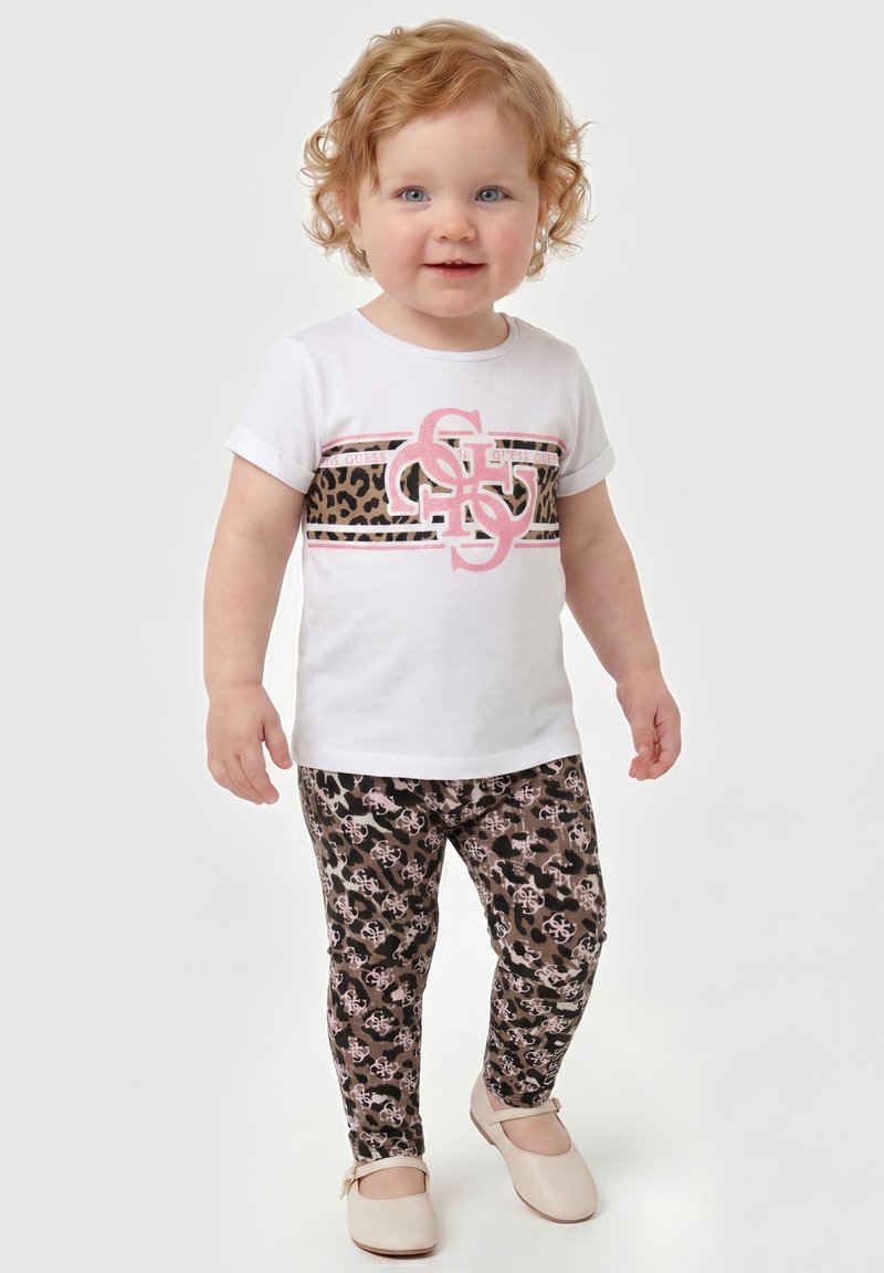 Tout-petit aux cheveux bouclés portant un t-shirt blanc avec imprimé léopard et logo, leggings imprimé léopard, et chaussures beiges, debout sur fond blanc.