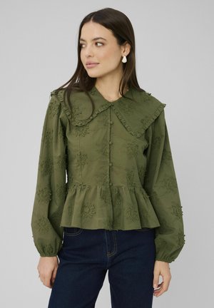 Femme aux longs cheveux foncés portant un chemisier vert olive brodé avec un grand col et un ourlet à volants, associé à un jean bleu foncé.