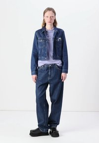 Giacca di jeans con un taglio corto, dotata di tasche frontali e bottoni metallici, abbinata a una t-shirt viola chiara e jeans a vestibilità ampia. Scarpe nere.