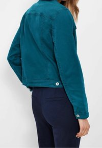 Tyrkysová džínová bunda s krátkým střihem, zapínáním na knoflíky a hladkou texturou. Stylizována s námořnickými kalhotami pro kontrast.