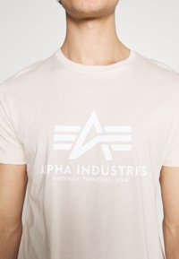 Beige bavlněné tričko s velkým bílým grafickým designem, na kterém je písmeno 'A' a tři pruhy, s textem "ALPHA INDUSTRIES, KNOXVILLE, TENNESSEE, U.S.A."