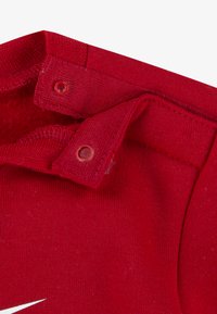 Ενδύματα από κόκκινο βαμβάκι  με κούμπωμα πατιλέτας και απαλό ύφασμα. Ορατές ραφές και μεγάλο λευκό λογότυπο. Στρογγυλεμένες άκρες και λείο φινίρισμα.