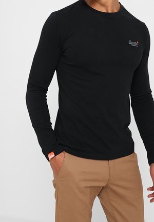 Man draagt een nauwsluitend zwart shirt met lange mouwen, klein logo op de borst en een oranje label aan de pols, gecombineerd met lichtbruine broek.