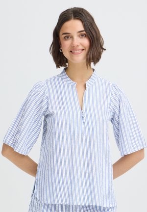 Femme souriante aux cheveux bruns courts portant un chemisier à rayures verticales bleues et blanches avec des manches mi-longues, debout devant un fond uni.