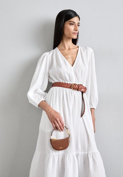Robe blanche fluide avec un décolleté en V plongeant et des manches bouffantes, cintrée à la taille par une ceinture en cuir tressé marron. Tient un petit sac à main marron.