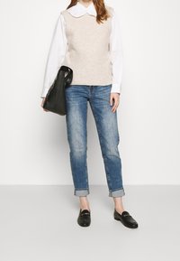 Béžová pletená vesta přes bílou košili s límečkem, doplněná o vyhrnuté modré džíny a černé mokasíny. Drží černou kabelku.