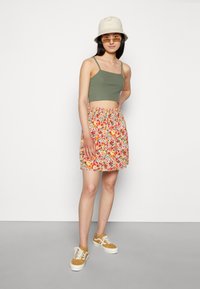 Top verde, saia skater com padrão floral em rosa, laranja e amarelo, combinada com ténis amarelos e um chapéu tipo balde bege.