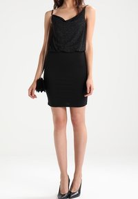 Czarny top bez rękawów o teksturowanym wykończeniu i głębokim dekolcie, w połączeniu z dopasowaną czarną mini spódnicą i czarnymi szpilkami.
