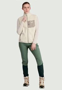 Fleeceweste in Off-White mit einer grauen Brusttasche, kombiniert mit grüner Hose mit dunkelgrünen Bündchen und beigen Wanderschuhen.