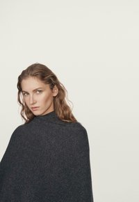 Žena s dlouhými vlnitými vlasy, oblečená v tmavě šedém pleteném ponču s vysokým límcem na jednobarevném světlém pozadí.