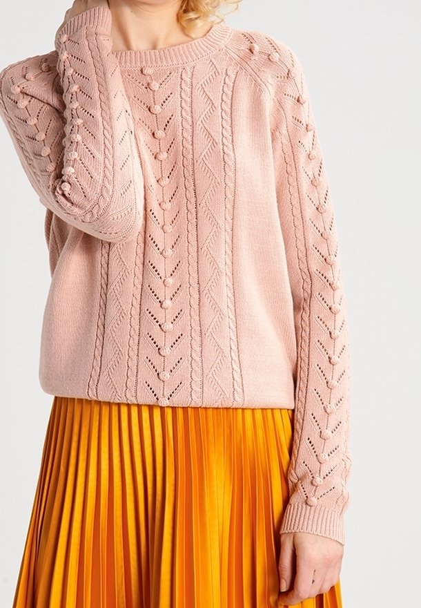 Femme portant un pull en maille texturée rose clair avec des détails en pompons et une jupe plissée orange vif sur un fond uni.