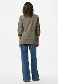 Žena s kovrčavom smešnom kosom koja nosi bež i crnu blazer s uzorkom ptičjeg zuba, plave široke traperice i smeđe cipele, okrenuta leđima.