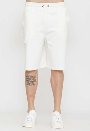 Pantaloncini bianchi al ginocchio con coulisse indossati da una persona con un tatuaggio in corsivo sul polpaccio sinistro e sneakers bianche.