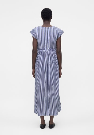 Γυναίκα που στέκεται με την πλάτη προς τα εκεί, φορώντας ένα midi φόρεμα με κάθετες μπλε και λευκές ρίγες και μαύρα παπούτσια slip-on σε μονόχρωμο φόντο.