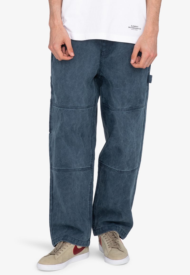 Weite blaue Baumwollhosen mit einer ausgeblichenen Textur, die sichtbare Nahtdetails und Seitentaschen aufweisen, kombiniert mit beigen Sneakers.